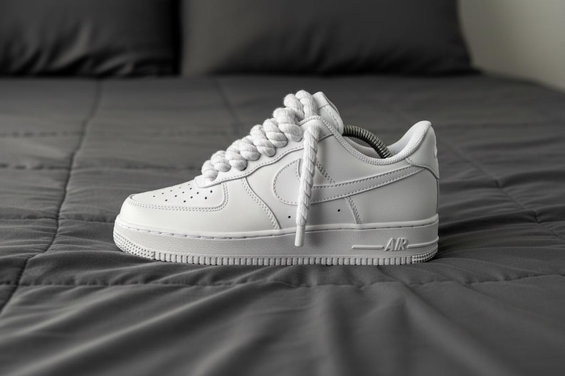 May include: A white leather sneaker with a classic design, featuring a perforated toe box, a prominent Swoosh logo, and thick white laces. The shoe is set against a gray background.