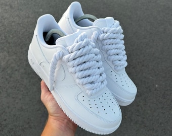 May include: A pair of white trainers featuring thick, rope-style laces. The shoes have a classic design with a perforated toe box and a white sole. The trainers are held up against a grey background.