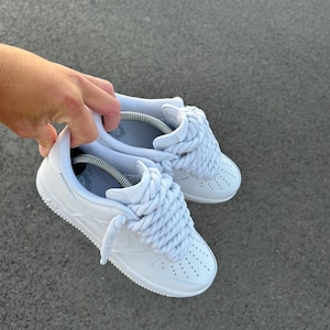 May include: White sneakers with thick, light blue rope laces. The shoes have a classic design with a perforated toe box and a white sole. The sneakers are held up against a gray background.