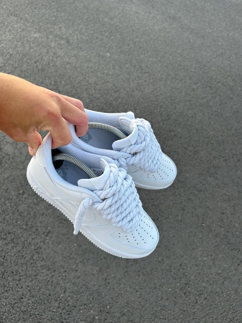 May include: White sneakers with thick, light blue rope laces. The shoes have a classic design with a perforated toe box and a white sole. The sneakers are held up against a gray background.