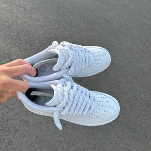 May include: A pair of white sneakers with thick, braided white laces. The shoes have a classic design with a perforated toe box and are displayed against a gray surface.