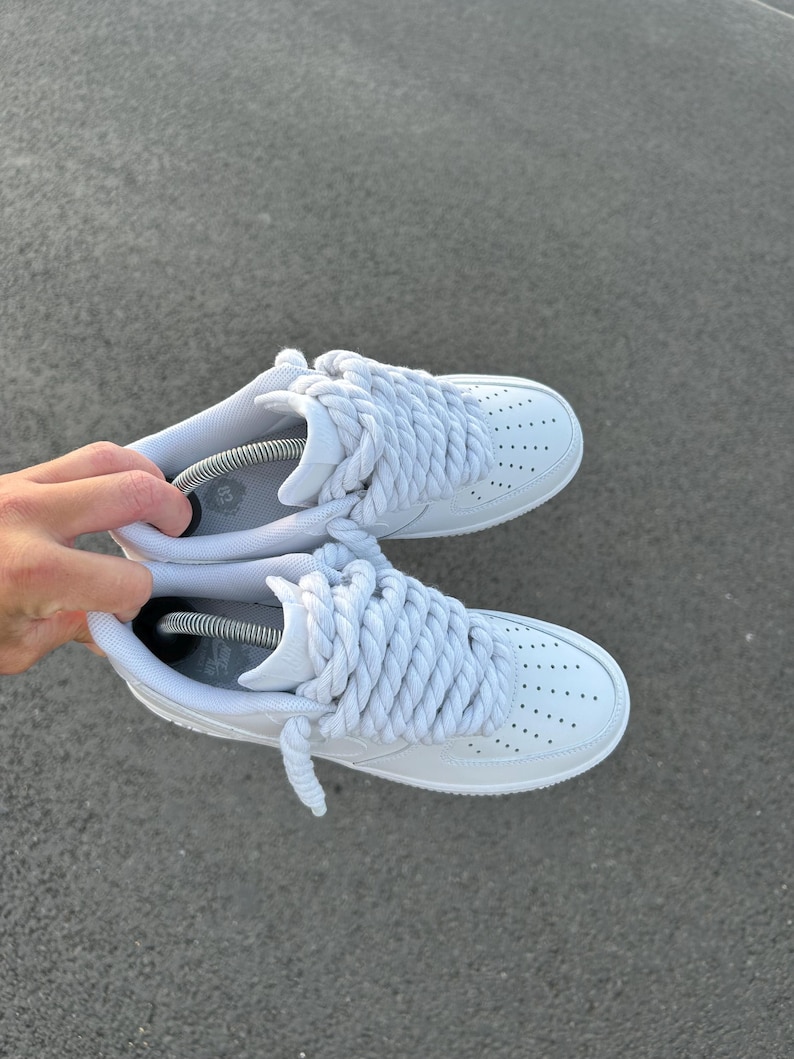 May include: A pair of white sneakers with thick, braided white laces. The shoes have a classic design with a perforated toe box and are displayed against a gray surface.