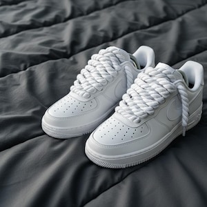 May include: A pair of white sneakers with thick, rope-like laces. The shoes have a classic design with a low profile, visible stitching, and small perforations on the toe box. The sneakers are set against a dark gray, textured background.
