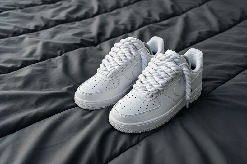 May include: A pair of white sneakers with thick, rope-like laces. The shoes have a classic design with a low profile, visible stitching, and small perforations on the toe box. The sneakers are set against a dark gray, textured background.