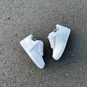 May include: A pair of white sneakers with thick, white laces. The shoes are made of a smooth material and have a classic design. The sneakers are set against a dark gray asphalt background.