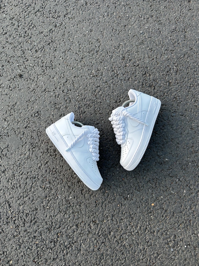 May include: A pair of white sneakers with thick, white laces. The shoes are made of a smooth material and have a classic design. The sneakers are set against a dark gray asphalt background.