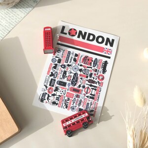 Può includere: Una collezione di souvenir a tema Londra. Include una cabina telefonica rossa, un autobus a due piani e un foglio con la scritta "LONDON" e varie icone londinesi. L'autobus ha la scritta "I LOVE LONDON".