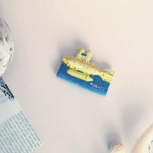 Può includere: Un magnete souvenir giallo e blu con un design a forma di barca e la scritta "MONACO" stampata sopra. Il magnete è rettangolare e sembra essere fatto di resina. È un oggetto a tema viaggio.