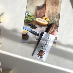 Op de afbeelding: Een stilleven met een baguette-verpakking met de tekst "Baguette Saveurs de France". De afbeelding bevat ook een watermeloen, cantaloupe, mes, snijplanken en een glazen fles. De scène speelt zich af op een wit oppervlak.