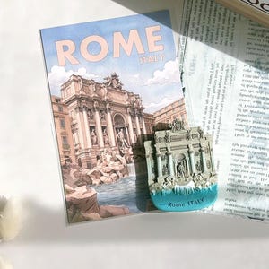 Peut inclure: Une carte postale et un aimant représentant la fontaine de Trevi à Rome. La carte postale affiche "ROME ITALY" en rose clair. L'aimant est une réplique 3D de la fontaine avec une base bleue et le texte "Rome ITALY".