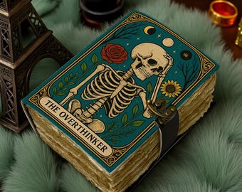 May include: A decorative journal with a teal cover featuring a skeleton illustration, a red rose, and the text "THE OVERTHINKER." The journal has aged-looking pages and a black leather strap closure. The cover has gold accents.