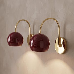 May include: Two wall-mounted lamps featuring glossy burgundy spherical shades and polished gold-tone curved arms. Each lamp has a circular gold-tone wall plate. The text GLOSSY BURGUNDY is visible in the upper left corner of the image.