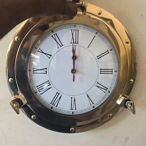 May include: A round wall clock with a polished brass porthole-style frame. The clock face is white with black Roman numerals and thin black hour and minute hands, along with a thin red second hand. The frame features decorative metal bolts.