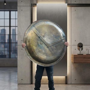 May include: A large, circular, metallic shield with a weathered, brass-toned finish and a prominent, raised V-shaped emblem across the center. The shield features a textured, uneven surface with visible signs of oxidation and metallic patina.