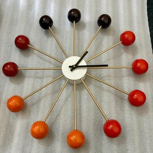Può includere: Un orologio da parete a raggiera con un centro circolare bianco e lancette nere. Dodici sottili raggi metallici color oro si irradiano verso l'esterno, ciascuno terminante con una sfera lucida in varie tonalità di marrone, rosso e arancione.