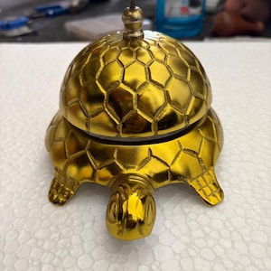 Pode incluir: Um sino dourado em forma de tartaruga com uma cúpula texturizada com padrão de favo de mel. O sino tem um pequeno topo em forma de cogumelo e é feito de um material brilhante e reflexivo. O sino foi projetado para ficar em uma superfície plana.