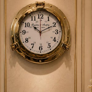 Könnte beinhalten: Eine runde Wanduhr mit einem polierten goldfarbenen Metallrahmen, der einem Schiffsbullauge ähnelt. Das weiße Zifferblatt zeigt schwarze arabische Ziffern von 1 bis 12, kleinere 13- bis 24-Stunden-Markierungen und den Text Franklin & Murphy EST 1868.