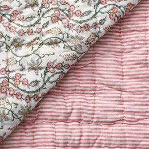 May include: A reversible quilted blanket. One side displays a floral design with pink flowers and green vines on a white backdrop. The other side has pink and white horizontal stripes. The blanket is suitable for a bed or sofa.