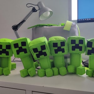 May include: Six green plush toys resembling Minecraft Creepers are arranged on a white surface. Each has a square head with a black pixelated face and a rectangular body with four blocky feet. The toys are in various poses.