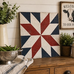 May include: A square wooden sign featuring a painted red, white, and blue star quilt pattern with a distressed finish. Beside it, a framed sign displays a black and white cow illustration with the text WELCOME TO OUR HOME in black lettering.