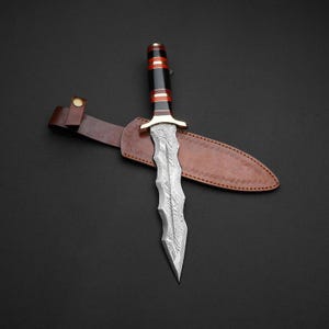 May include: A Damascus steel dagger with a decorative handle featuring black, red, and gold accents. The dagger rests on a brown leather sheath with a belt loop, set against a dark background.