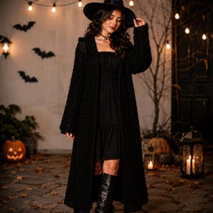 May include: A long, black, open-front crochet cardigan with long sleeves, worn over a black dress, paired with a wide-brimmed black hat and black boots. The garment features a textured, chunky knit pattern and a straight, floor-length silhouette.