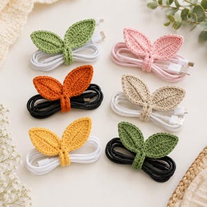 May include: Six coiled charging cables secured with crocheted leaf-shaped organizers. The set includes green, pink, orange, beige, yellow, and dark green leaf designs wrapped around white or black USB cables with standard connectors.