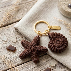 Puede incluir: Un llavero de metal dorado con dos dijes de ganchillo de color marrón oscuro, uno con forma de estrella de mar de cinco puntas y otro con forma de concha en espiral, con una pequeña etiqueta rectangular grabada con la palabra Handmade.