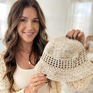 May include: A beige crochet bucket hat featuring a solid crown and a wide, open-weave lattice pattern around the mid-section, finished with a solid brim. The hat is constructed from a textured, natural-toned yarn with a consistent stitch pattern.