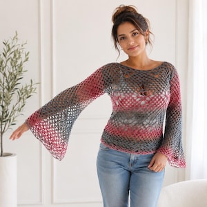 Puede incluir: Una blusa de ganchillo de manga larga con un patrón de malla abierta y un degradado de color que va desde el gris oscuro en el cuello hasta un rosa vibrante en los puños acampanados y el dobladillo, sobre una camiseta clara y vaqueros azules.
