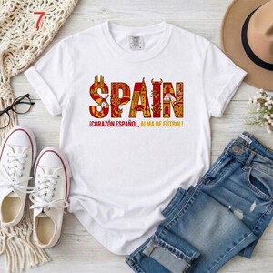 May include: A white short-sleeved t-shirt featuring the word SPAIN in large, red and orange letters filled with intricate illustrations of Spanish landmarks and symbols. Below the word, the text reads &iexcl;CORAZ&Oacute;N ESPA&Ntilde;OL, ALMA DE F&Uacute;TBOL!