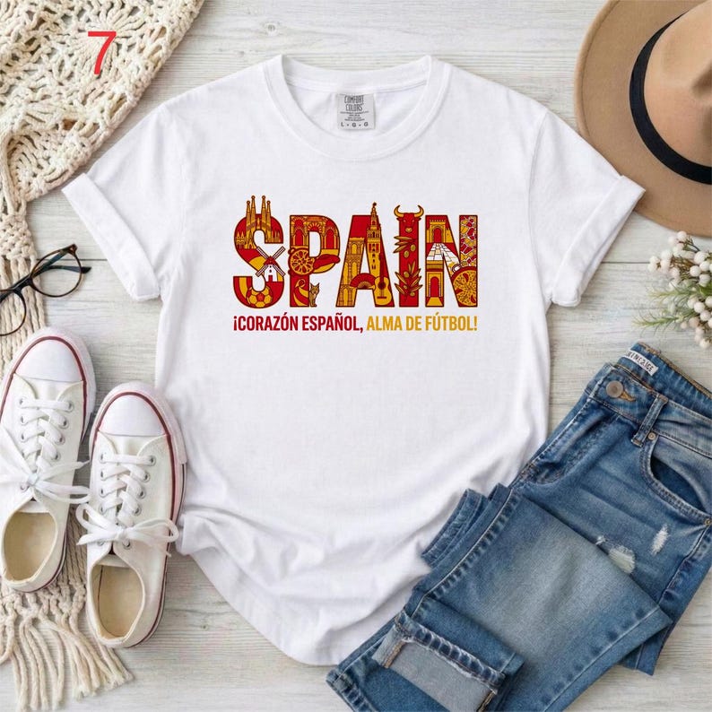 May include: A white short-sleeved t-shirt featuring the word SPAIN in large, red and orange letters filled with intricate illustrations of Spanish landmarks and symbols. Below the word, the text reads &iexcl;CORAZ&Oacute;N ESPA&Ntilde;OL, ALMA DE F&Uacute;TBOL!