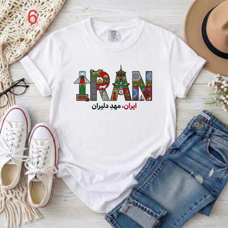 May include: A white short-sleeve t-shirt featuring a graphic print of the word IRAN in colorful, patterned letters filled with cultural motifs and landmarks. Below the graphic, the text reads Iran, Mahd-e Daliran in Persian script.
