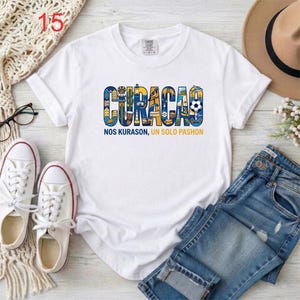 May include: A white short-sleeve t-shirt featuring a graphic of the word CURACAO in blue and yellow, filled with illustrations of local landmarks, a soccer ball, and a bird. Below the graphic is the text NOS KURASON, UN SOLO PASHON.