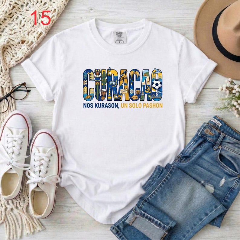 May include: A white short-sleeve t-shirt featuring a graphic of the word CURACAO in blue and yellow, filled with illustrations of local landmarks, a soccer ball, and a bird. Below the graphic is the text NOS KURASON, UN SOLO PASHON.