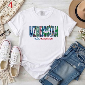 May include: A white short-sleeved t-shirt featuring the word UZBEKISTAN in large, multi-colored letters filled with intricate patterns and cultural motifs. Below the main text, the phrase OLĞA, O'ZBEKISTON! is printed in blue and red lettering.