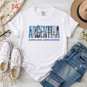 May include: A white short-sleeved t-shirt featuring the word ARGENTINA in large blue block letters filled with various small illustrations. Below the text, the phrase SIEMPRE JUNTOS, SIEMPRE ARGENTINA is printed in blue capital letters.