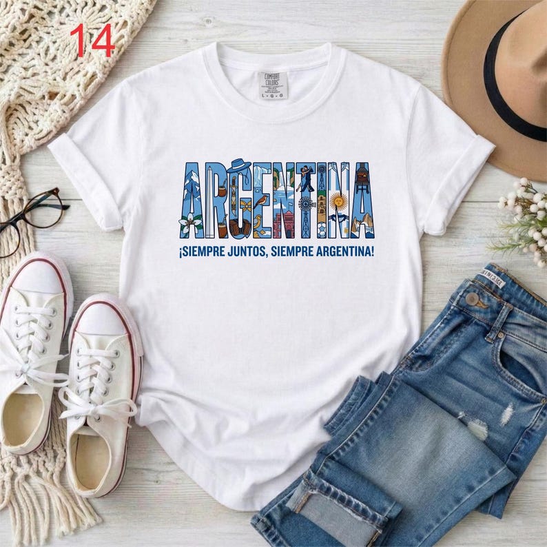 May include: A white short-sleeved t-shirt featuring the word ARGENTINA in large blue block letters filled with various small illustrations. Below the text, the phrase SIEMPRE JUNTOS, SIEMPRE ARGENTINA is printed in blue capital letters.