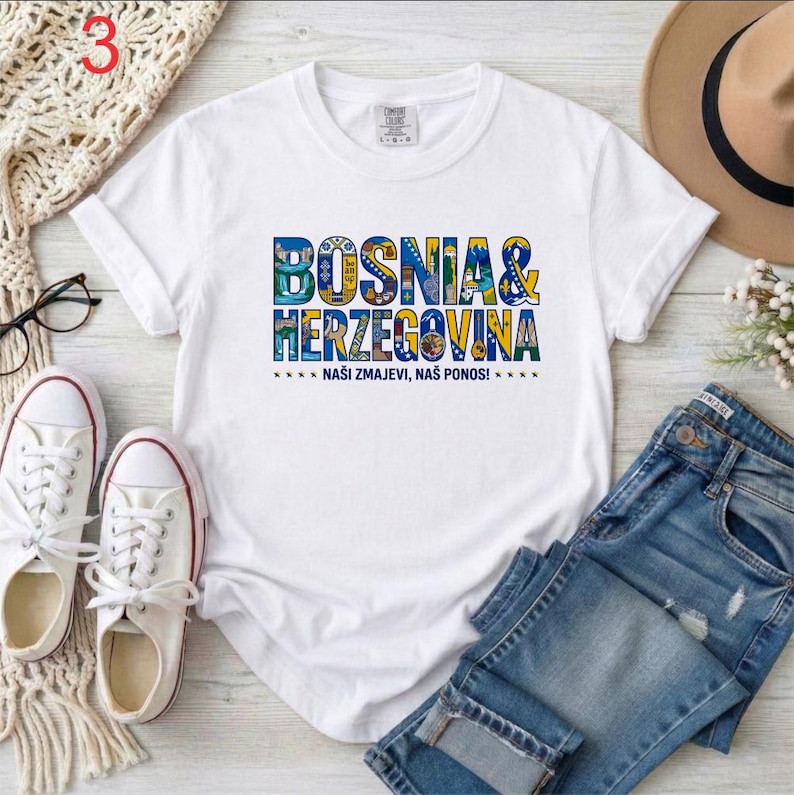 May include: A white short-sleeved t-shirt featuring the text BOSNIA & HERZEGOVINA in a stylized blue and yellow font with various small illustrations inside the letters, and the text NA&Scaron;I ZMAJEVI, NA&Scaron; PONOS! printed in smaller black letters below.