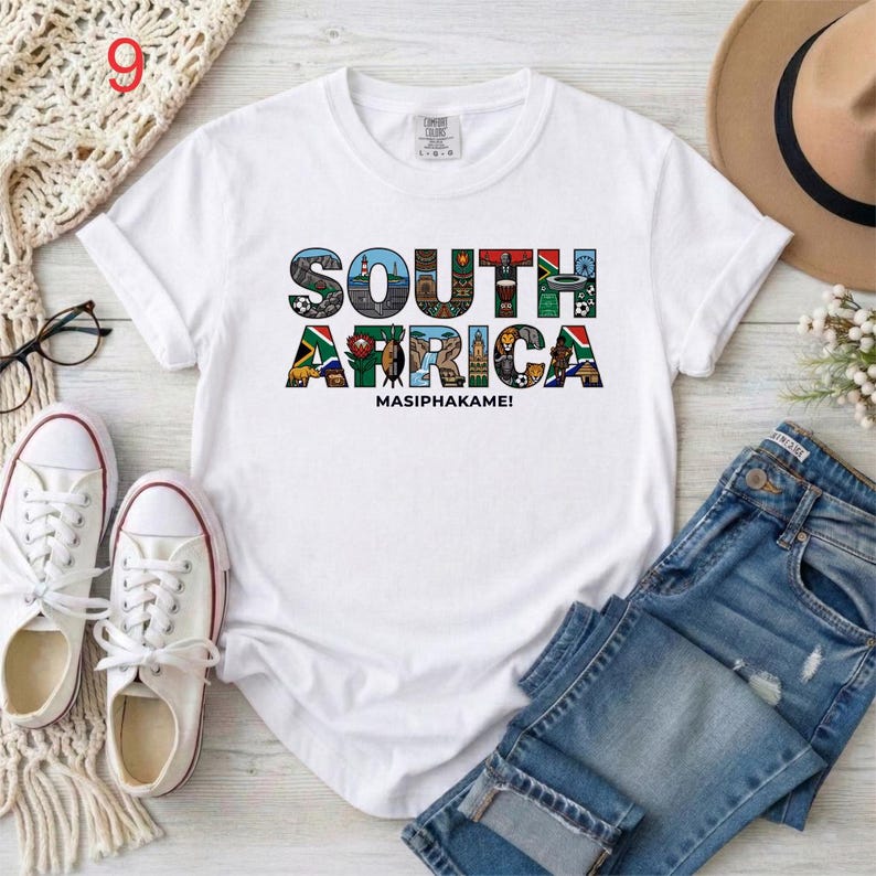 May include: A white short-sleeve t-shirt featuring the text SOUTH AFRICA in large block letters, with each letter filled with colorful illustrations of landmarks, wildlife, and cultural symbols, and the word MASIPHAKAME! printed below.