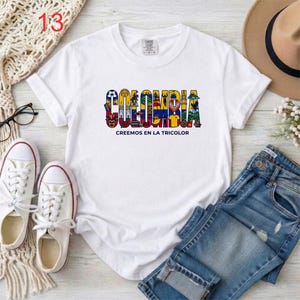 May include: A white short-sleeved t-shirt featuring the word COLOMBIA in large, colorful, illustrated letters filled with various cultural symbols and the text CREEMOS EN LA TRICOLOR printed in black underneath the main graphic design.
