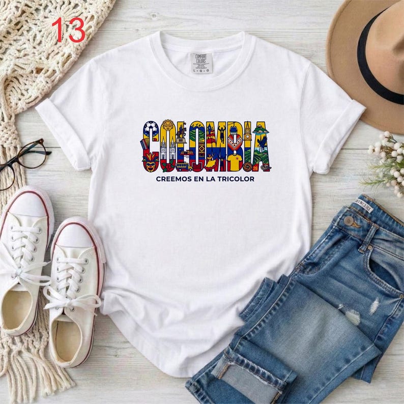 May include: A white short-sleeved t-shirt featuring the word COLOMBIA in large, colorful, illustrated letters filled with various cultural symbols and the text CREEMOS EN LA TRICOLOR printed in black underneath the main graphic design.
