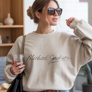 May include: A beige sweatshirt with the name "Michael Jackson" in a cursive font. The sweatshirt is a neutral color and has a crew neck. The image shows a person wearing the sweatshirt.
