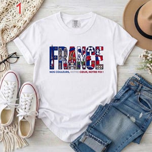 May include: A white short-sleeve t-shirt featuring the word FRANCE in large block letters filled with colorful illustrations of French landmarks and symbols, with the text NOS COULEURS, NOTRE C&OElig;UR, NOTRE FOI! printed in smaller letters below.