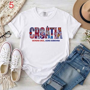 May include: A white short-sleeved t-shirt featuring the word CROATIA in large, stylized letters filled with red, white, and blue patterns. Below the word, the text reads VATRENO SRCE, JEDNA DOMOVINA in smaller blue and red font on the chest.
