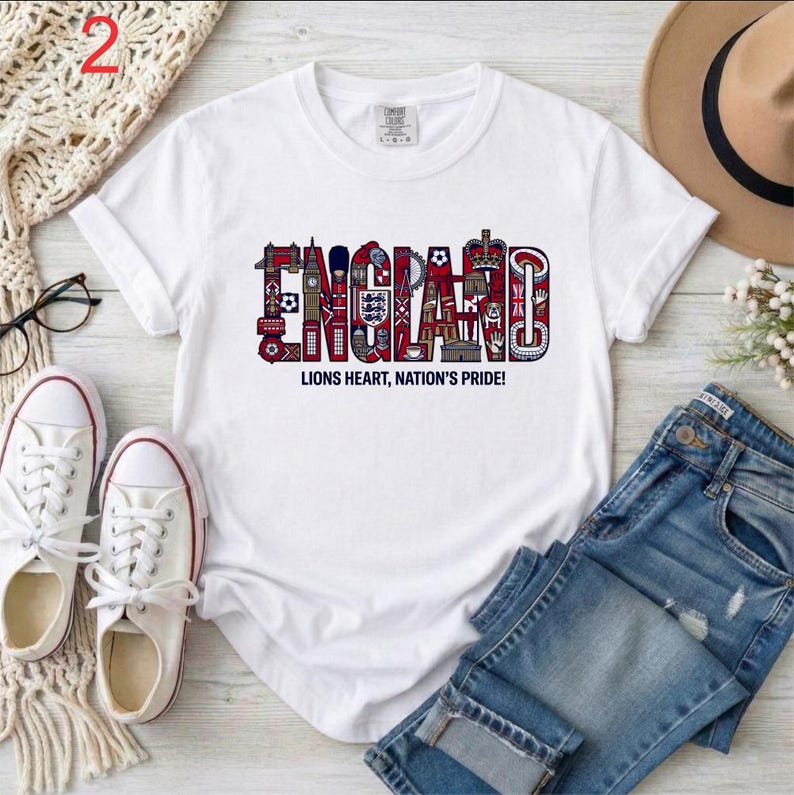 May include: A white short-sleeved t-shirt featuring the word ENGLAND in large, stylized letters filled with red, white, and blue illustrations of British landmarks and symbols, with the text LIONS HEART, NATION'S PRIDE! printed underneath.