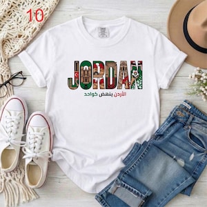 May include: A white short-sleeved t-shirt featuring the word JORDAN in large block letters, with each letter filled with colorful illustrations of Jordanian landmarks and cultural symbols, and Arabic text printed in green and red below.