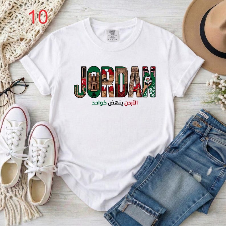 May include: A white short-sleeved t-shirt featuring the word JORDAN in large block letters, with each letter filled with colorful illustrations of Jordanian landmarks and cultural symbols, and Arabic text printed in green and red below.