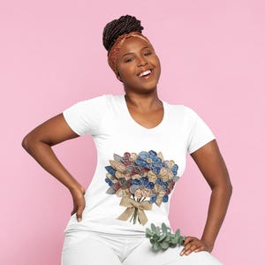 Puede incluir: Camiseta blanca con un diseño de ramo floral. El ramo incluye flores de tela azules, rojas y beige con un lazo de arpillera. La persona lleva un pañuelo en la cabeza estampado.