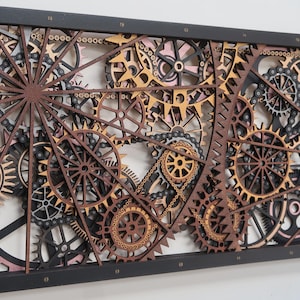 Puede incluir: Arte mural de madera que presenta una compleja disposición de engranajes, cadenas y piñones en varios tamaños y colores. Los engranajes son principalmente marrones, negros y beige, dentro de un marco rectangular negro. Estética steampunk.
